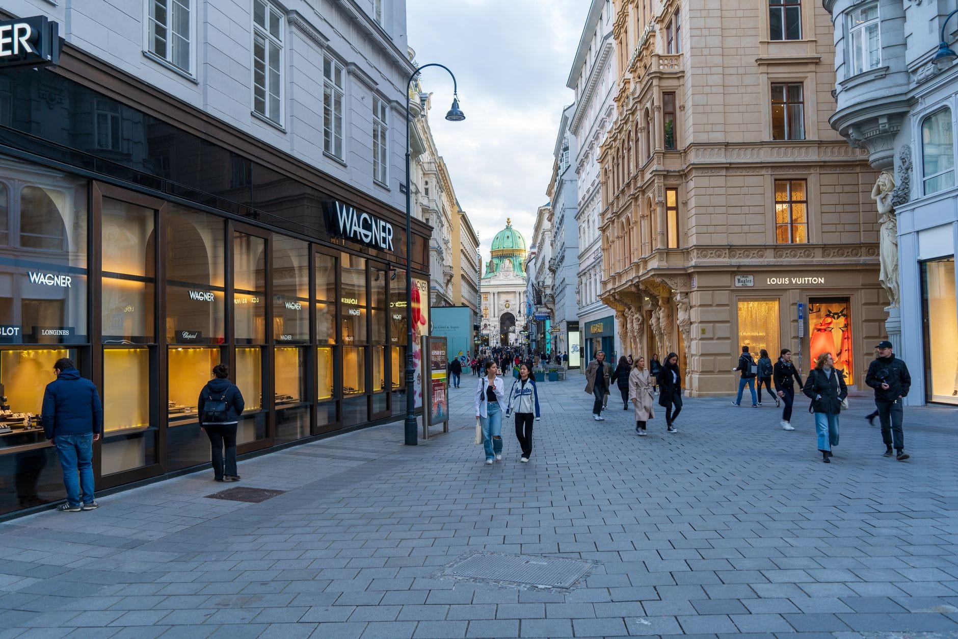 Fußgängerzone in Wiens historischer Innerer Stadt mit Passanten, eleganten Gebäuden und der grün bekuppelten Hofburg im Hintergrund.