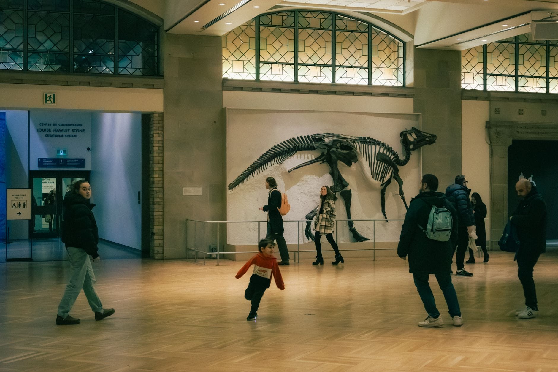 Familien und Kinder erkunden einen großen Museumssaal mit einem ausgestellten Dinosaurierskelett und hohen Bogenfenstern.