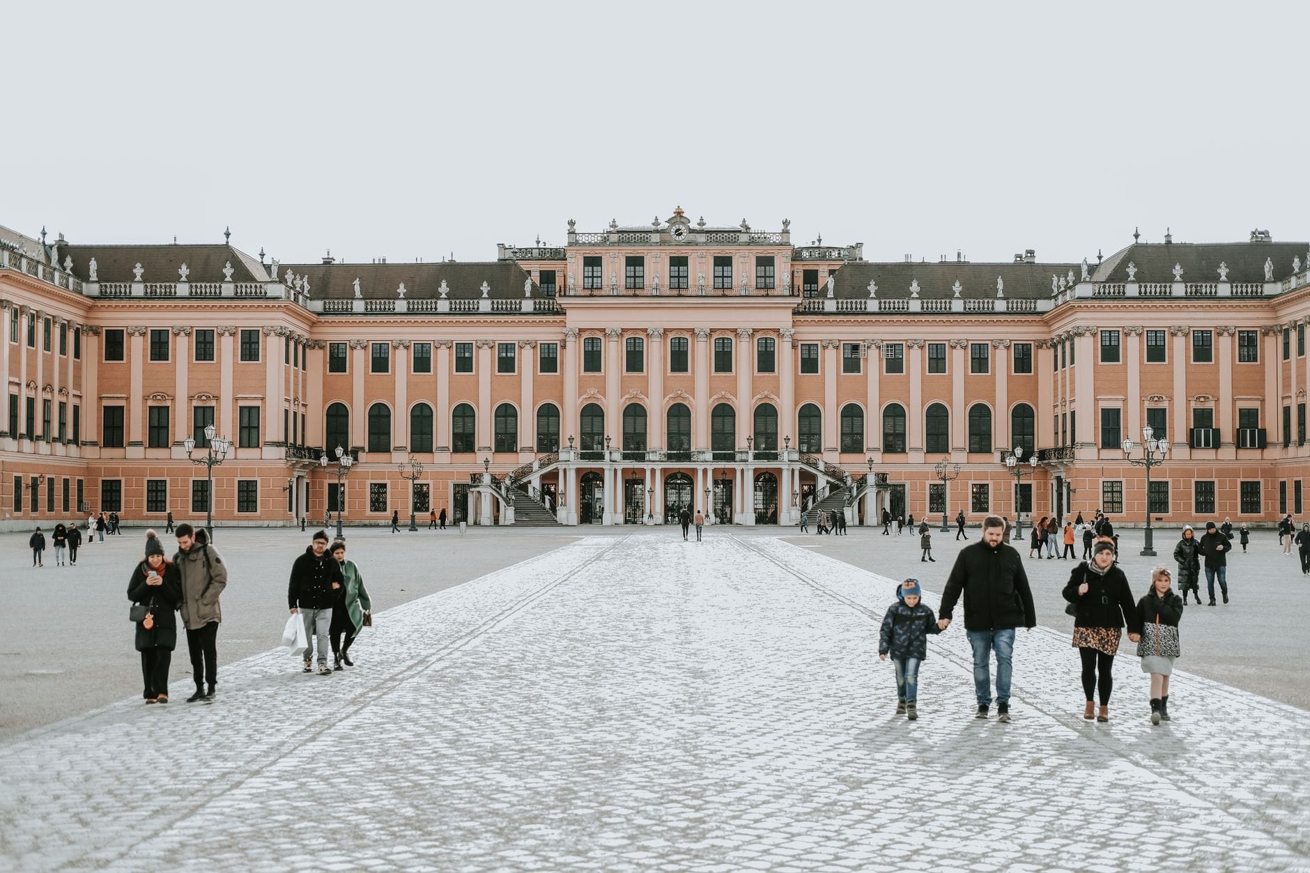 Familien mit Kindern spazieren bei strahlendem Sonnenschein vor dem imposanten Schloss Schönbrunn in Wien – eine einladende Atmosphäre für den Familienurlaub.