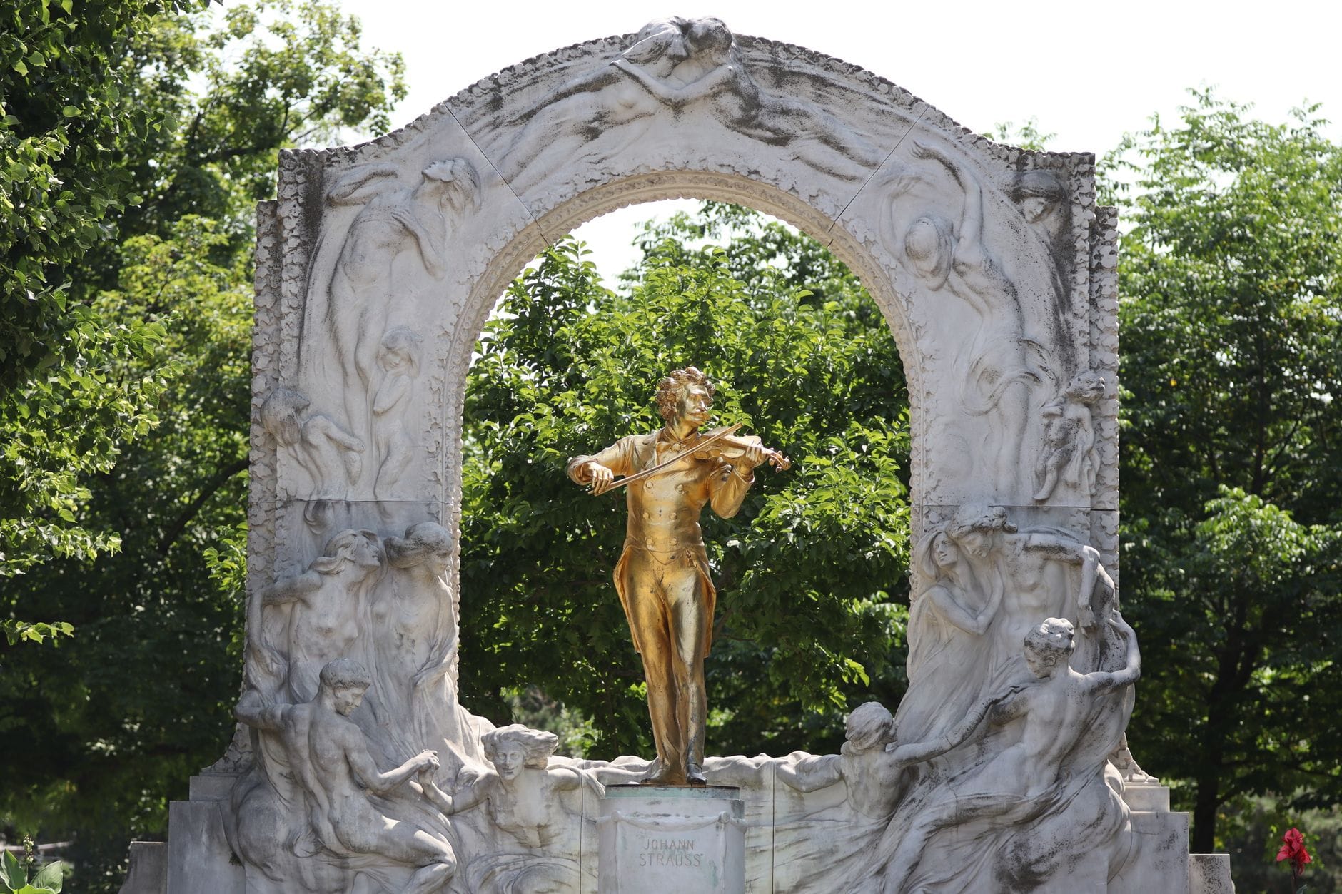 Berühmtes goldenes Johann-Strauss-Denkmal im Wiener Stadtpark, umgeben von grünen Bäumen und einem gemeißelten Marmorbogen.