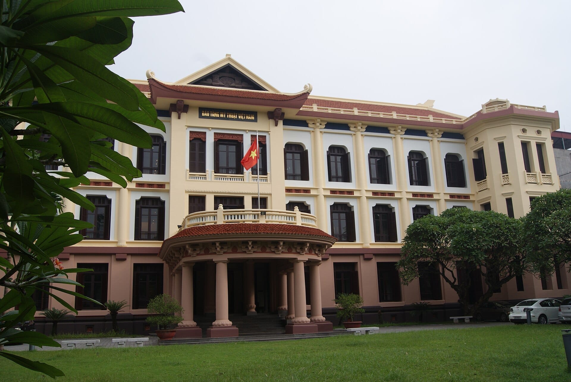 Front view of the Vietnam Fine Arts Museum in Hanoi, showcasing its French colonial architecture, Vietnamese flag, and green garden area on a cloudy day.