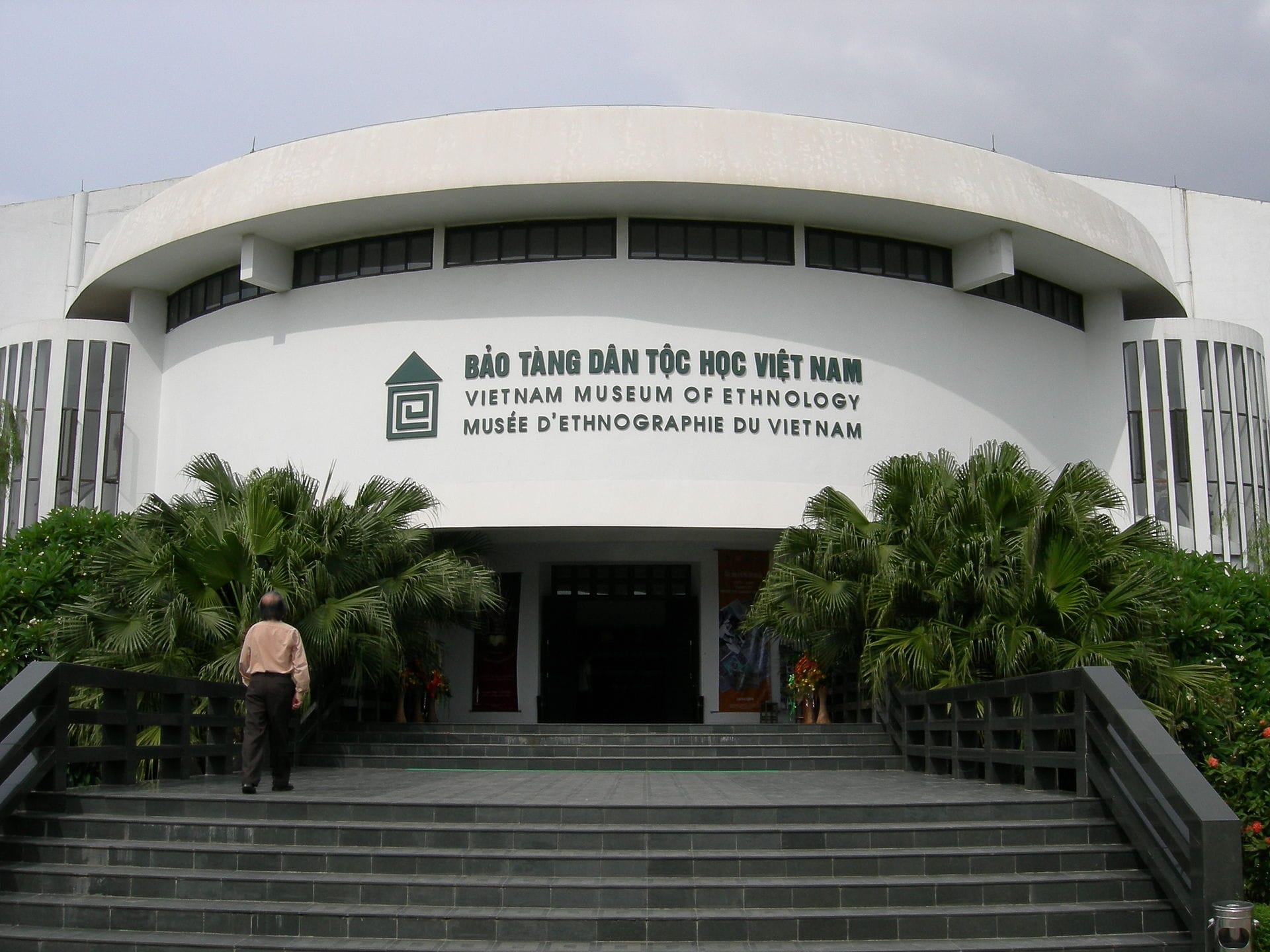Front entrance of the Vietnam Museum of Ethnology in Hanoi, with wide steps, lush green plants, and a person approaching the doorway.