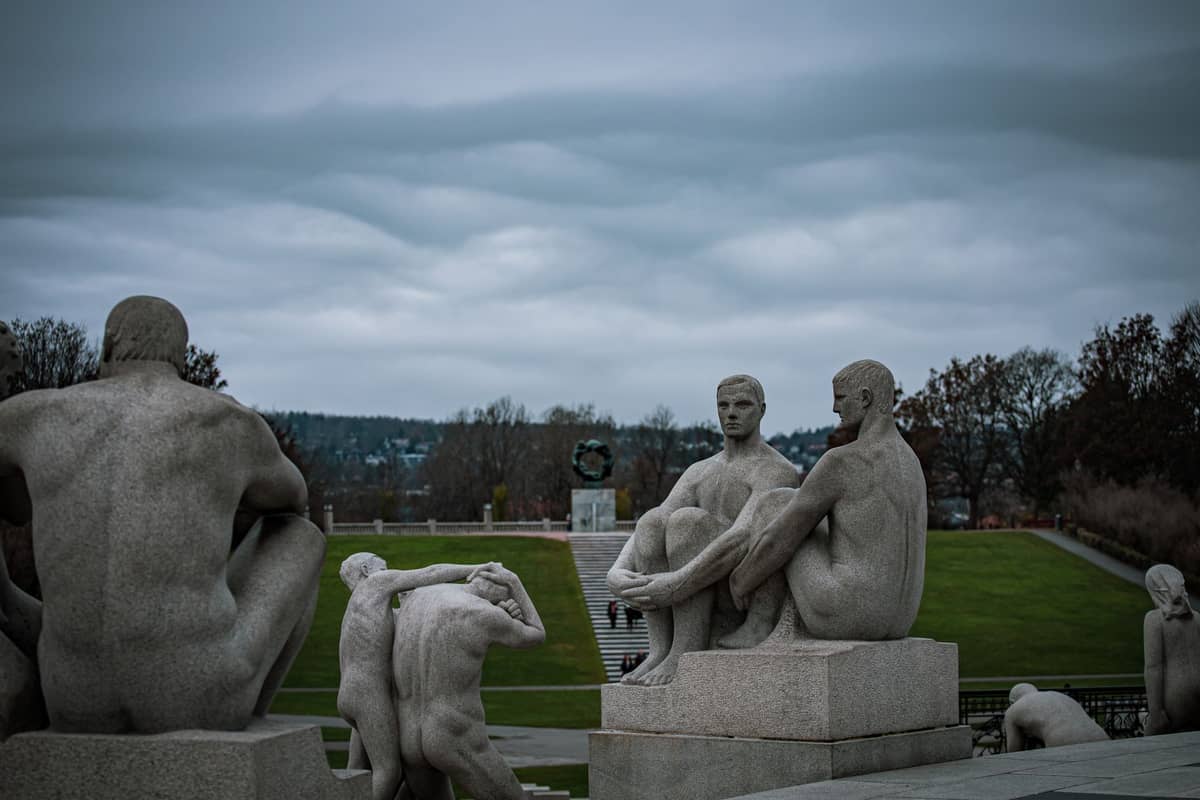ประติมากรรมหินแกรนิตรูปมนุษย์ในสวนประติมากรรม Vigeland ภายใต้ท้องฟ้าครึ้ม ล้อมรอบด้วยสนามหญ้าสีเขียวและต้นไม้ในระยะไกลที่เมือง Oslo ประเทศนอร์เวย์