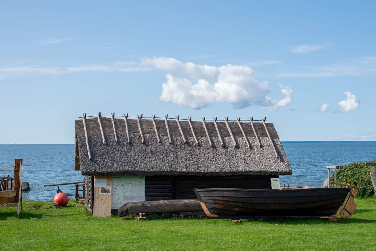 Traditionelles Bootshaus mit Reetdach und altem Holzfischerboot auf der Wiese davor, vor blauem Himmel und der Ostsee im Freilichtmuseum Viimsi.