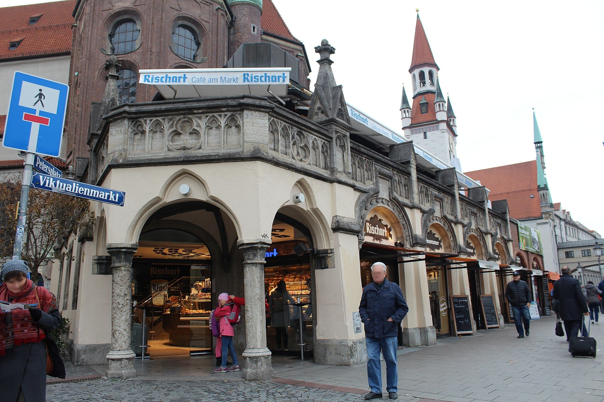 ภาพมุมถนนของทางเข้าตลาด Viktualienmarkt มิวนิก เห็นร้านเบเกอรี่ ผู้คน และป้าย Viktualienmarkt กับหอคอยอยู่พื้นหลัง