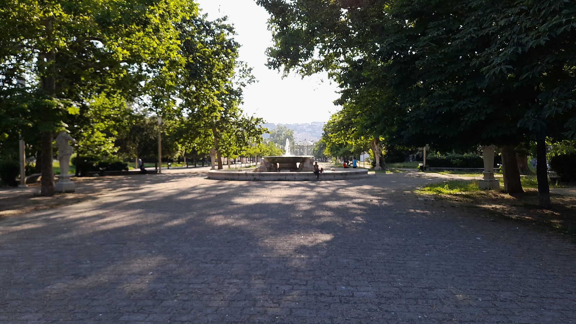 Scena soleggiata nel parco con una fontana centrale, viali alberati, panchine e persone che si rilassano alla Villa Comunale di Napoli.