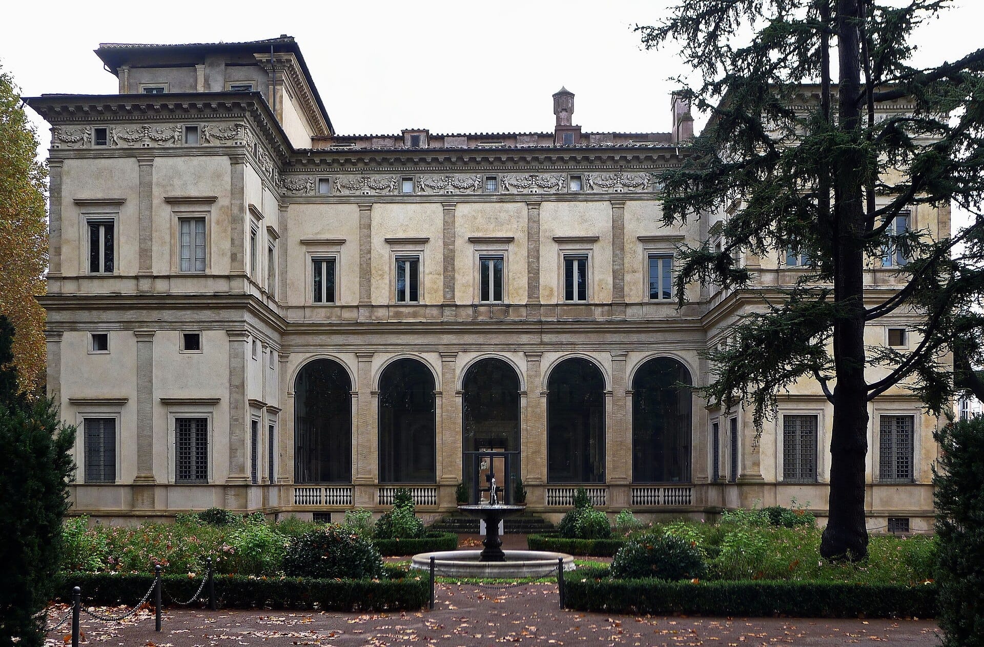 Front view of Villa Farnesina in Rome, featuring Renaissance architecture, large arched windows, manicured garden, and a central fountain under an overcast sky.