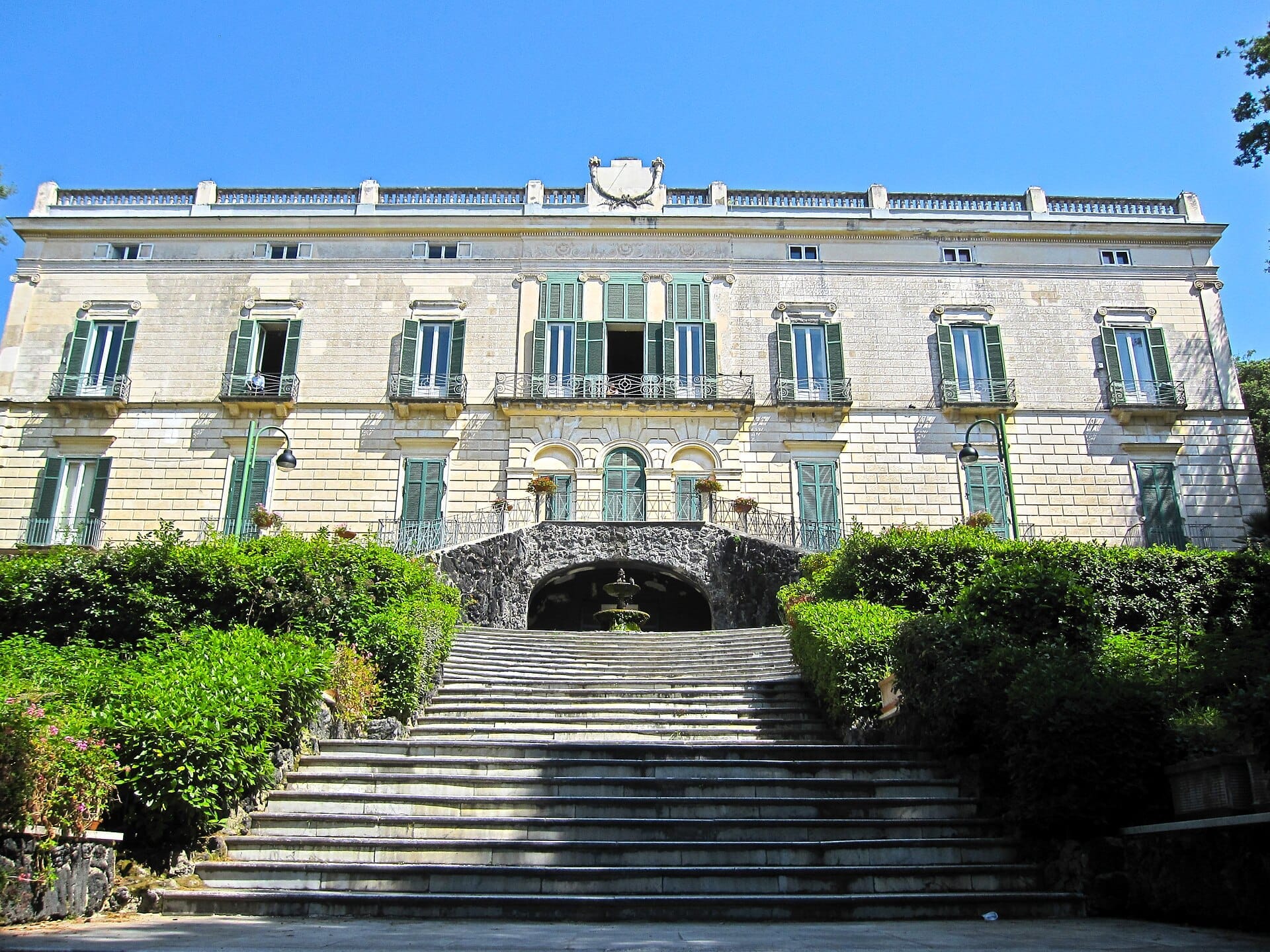 Una larga scalinata in pietra sale attraverso una vegetazione lussureggiante verso la neoclassica Villa Floridiana con le persiane verdi e il cielo blu sullo sfondo.