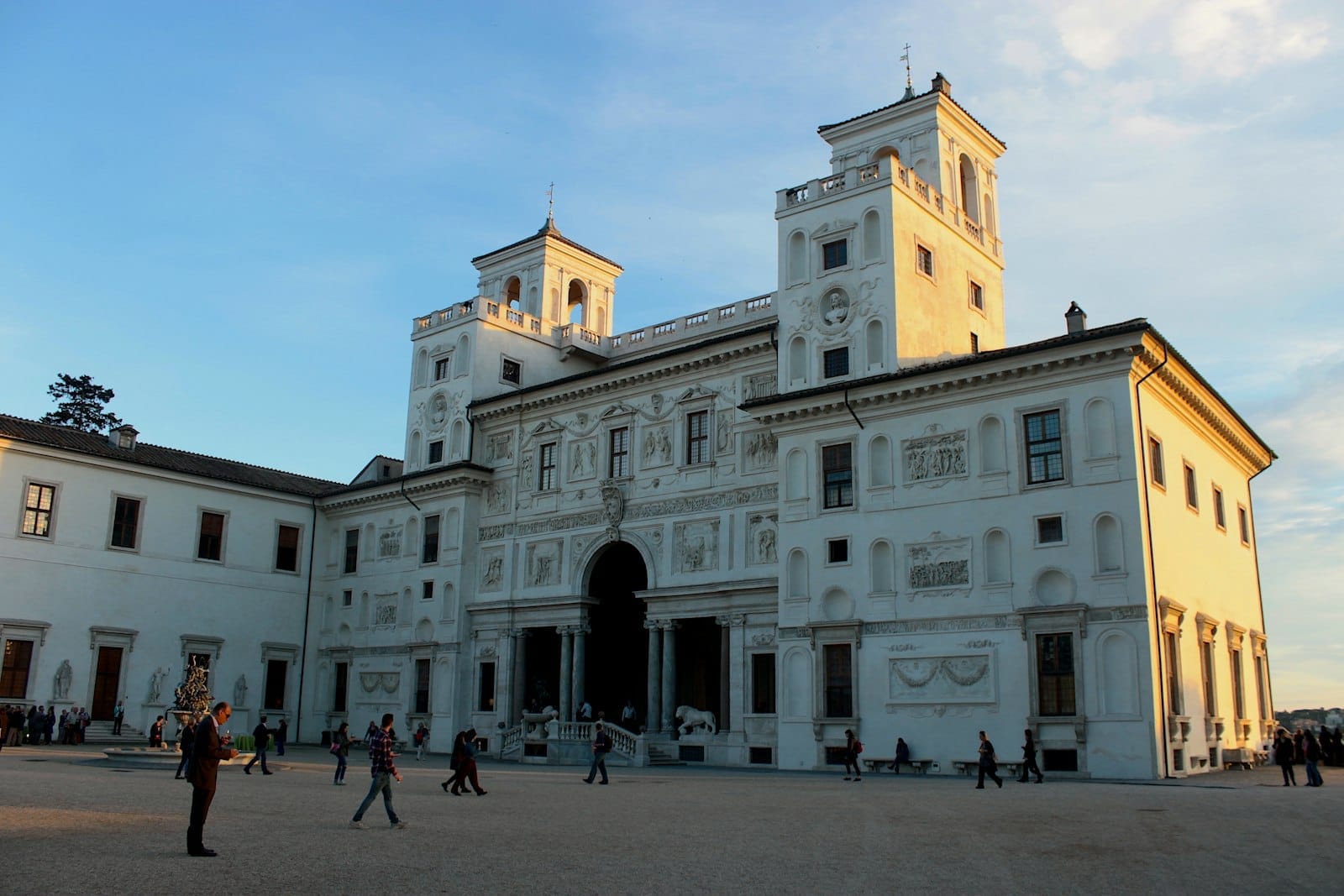 Facade of Villa Medici in Rome at sunset, with detailed stone carvings, twin towers, and visitors walking in the spacious courtyard.
