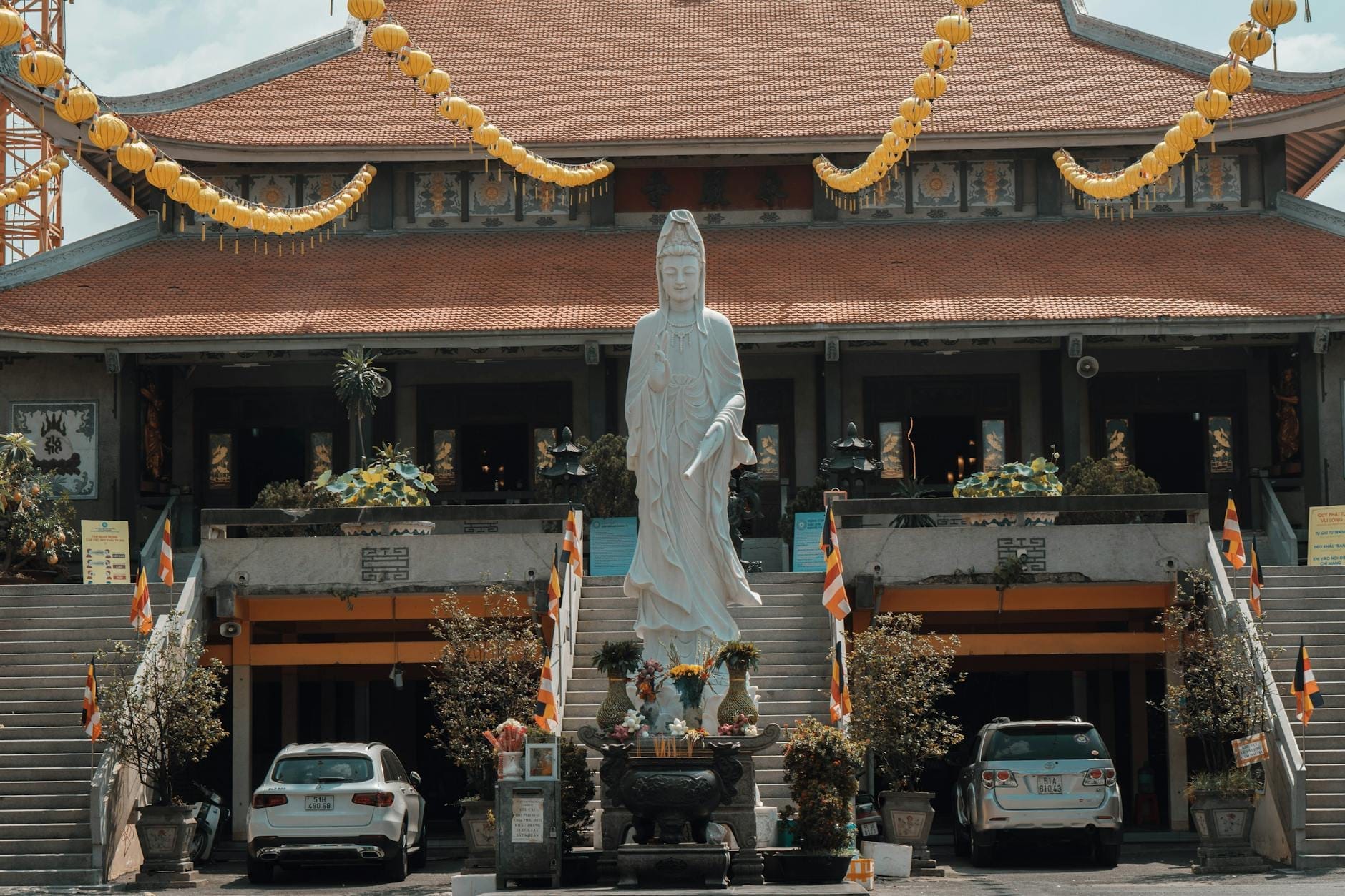 Vista frontal de la Pagoda Vĩnh Nghiêm con una gran estatua blanca, faroles amarillos y coches aparcados bajo la luz del día.