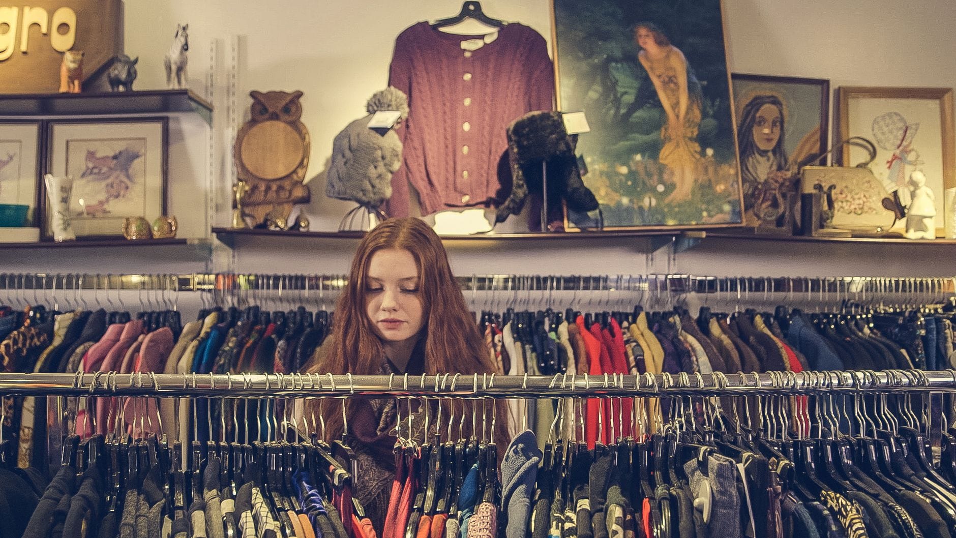 Young woman browsing through racks of colorful vintage clothing in a well-decorated boutique filled with art and retro items.