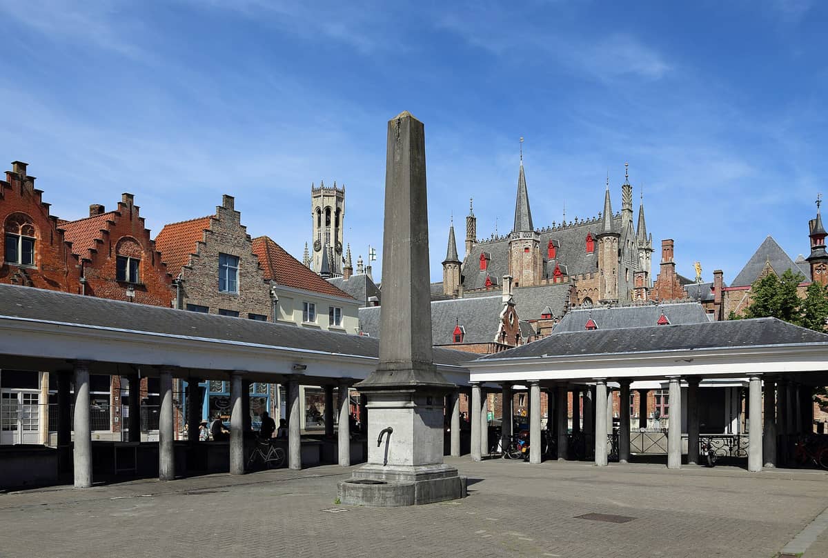 The covered open-air fish market (Vismarkt) in Bruges, with its colonnade and historic gabled buildings visible in the background under a blue sky.