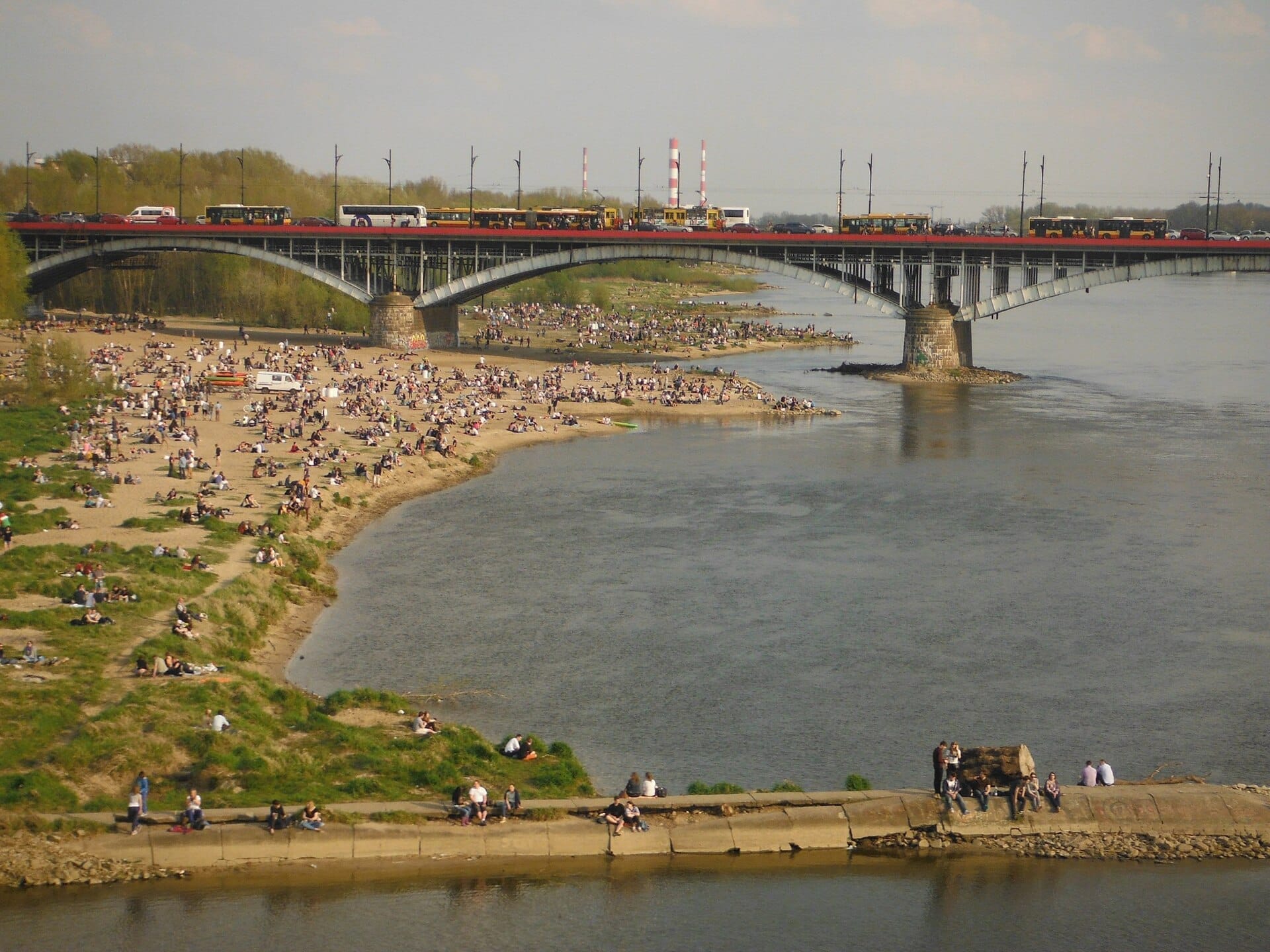 ฝูงชนพักผ่อนบนฝั่งทรายของหาด Vistula River ในกรุง Warsaw โดยมีสะพานและฉากหลังของเมืองใต้ท้องฟ้ามีเมฆบางส่วน