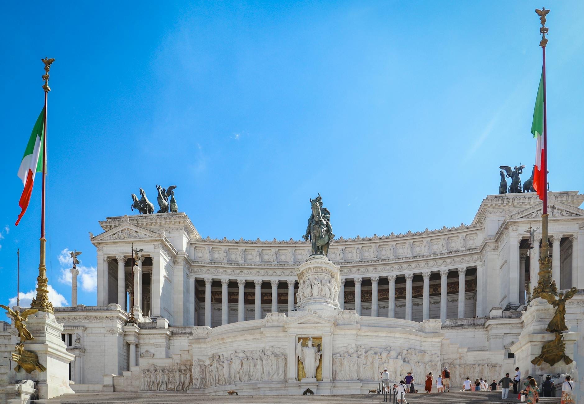 Front view of the Vittoriano monument in Rome with two Italian flags, blue sky, white marble and visitors on the steps.