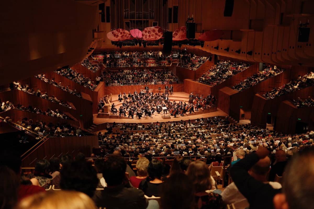 Blick vom Publikum im Inneren des Sydney Opera House während eines Live-Orchesterkonzerts, mit einem voll besetzten Saal und Musikern auf der Bühne.