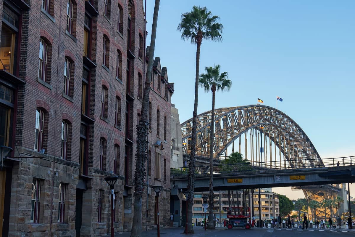 Blick auf die Sydney Harbour Bridge und historische Lagerhäuser im Viertel The Rocks an einem klaren Tag, mit Palmen und Spaziergängern in der Nähe.