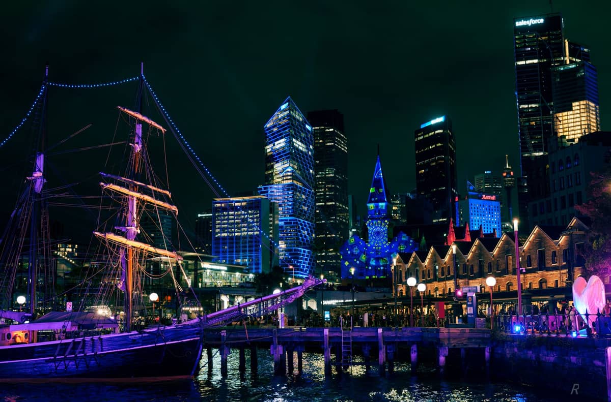 Skyline von Sydney bei Nacht während Vivid Sydney, mit Gebäuden und einem historischen Schiff, die am Ufer in bunten Lichtern erstrahlen.