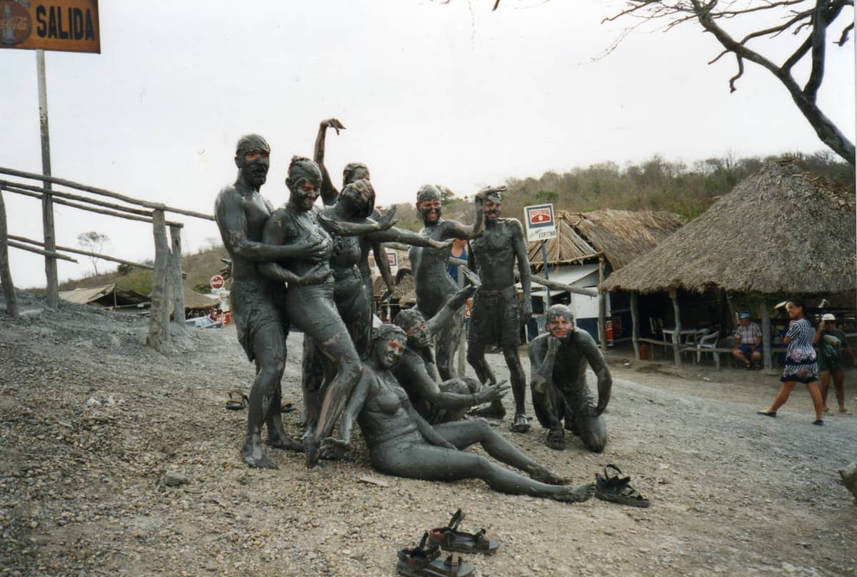A group of people covered in mud pose together outside Volcán de Lodo El Totumo, with rustic huts and hillside in the background.