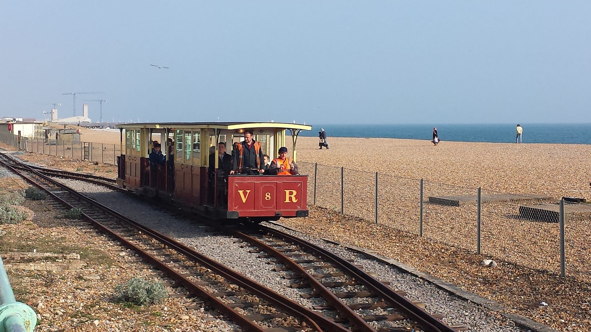 Volk's Electric Railway train travels along a seaside pebble beach in Brighton with passengers on board and clear blue skies overhead.