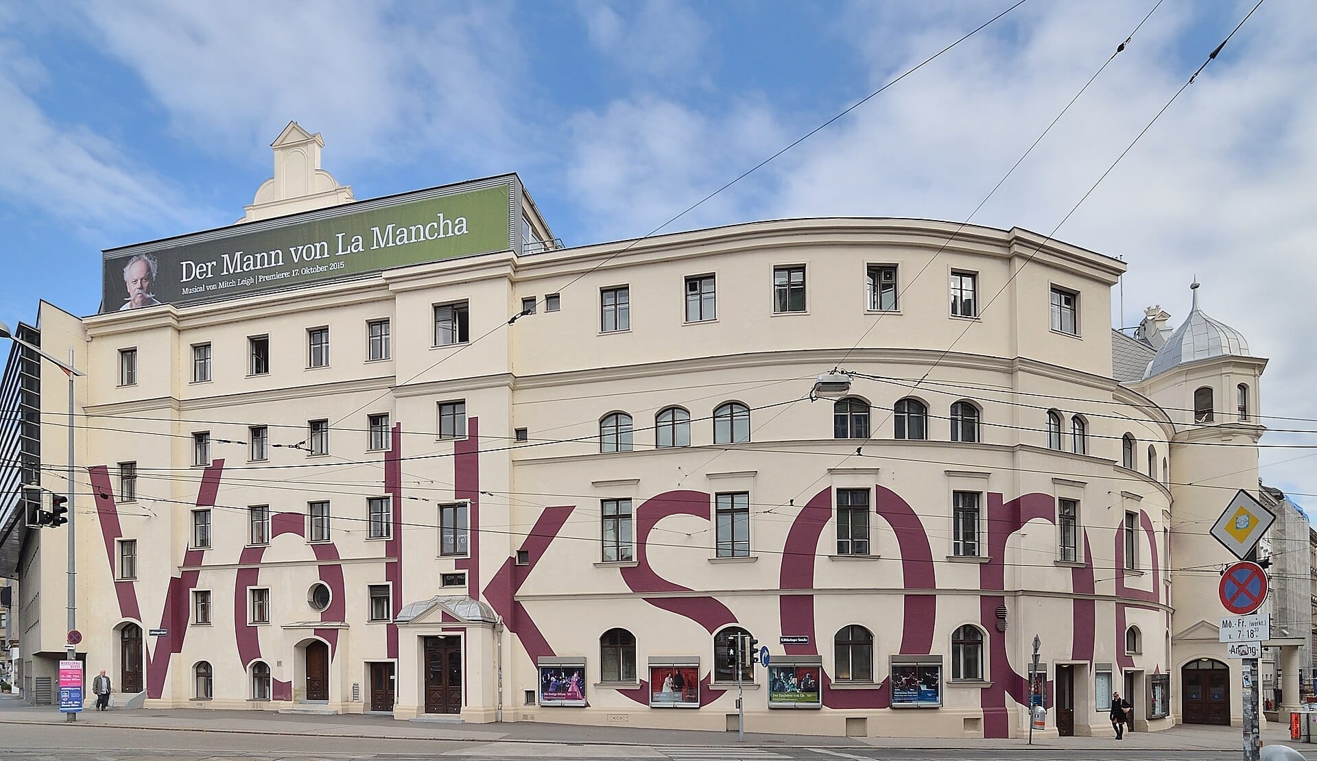 Frontansicht des Gebäudes der Volksoper Wien mit seinem großen modernen Schriftzug, cremefarbener Fassade und einem grünen Banner, das ein Musical bewirbt.