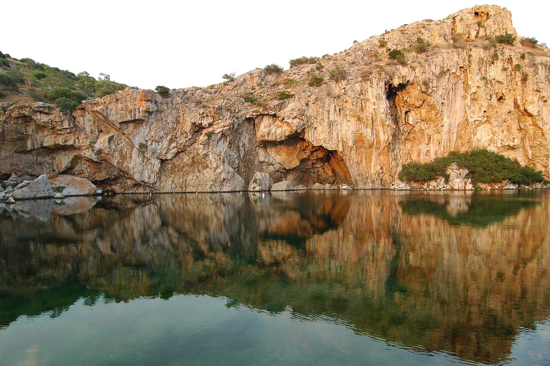 Il lago Vouliagmeni con le sue spettacolari scogliere rocciose riflesse in acque calme e cristalline sotto un cielo luminoso sulla Riviera ateniense.