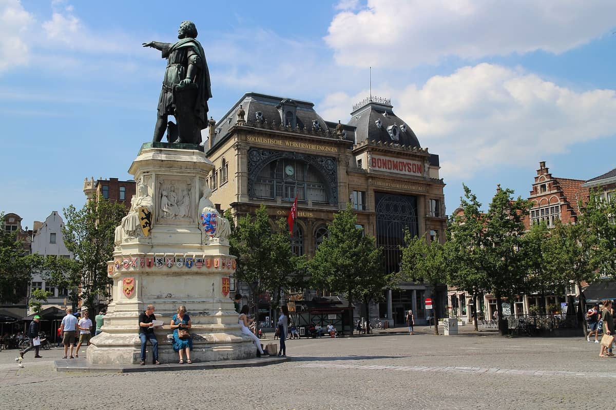 View of Vrijdagmarkt square in Ghent featuring the famous statue of Jacob van Artevelde, historic guild houses, and people sitting around the monument.
