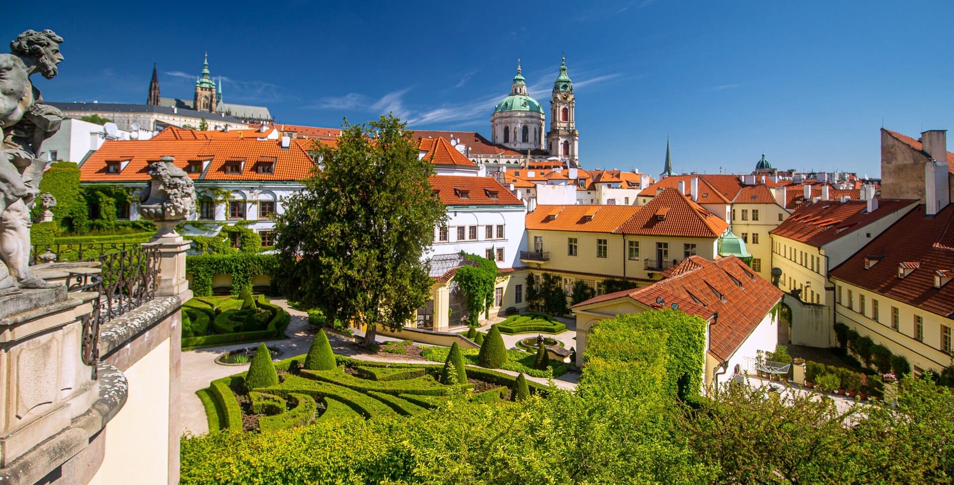 Prachtvoller barocker Vrtba-Garten mit gepflegten Hecken, Statuen und Gebäuden mit Terrakottadächern unter einem klaren blauen Himmel in Prag.