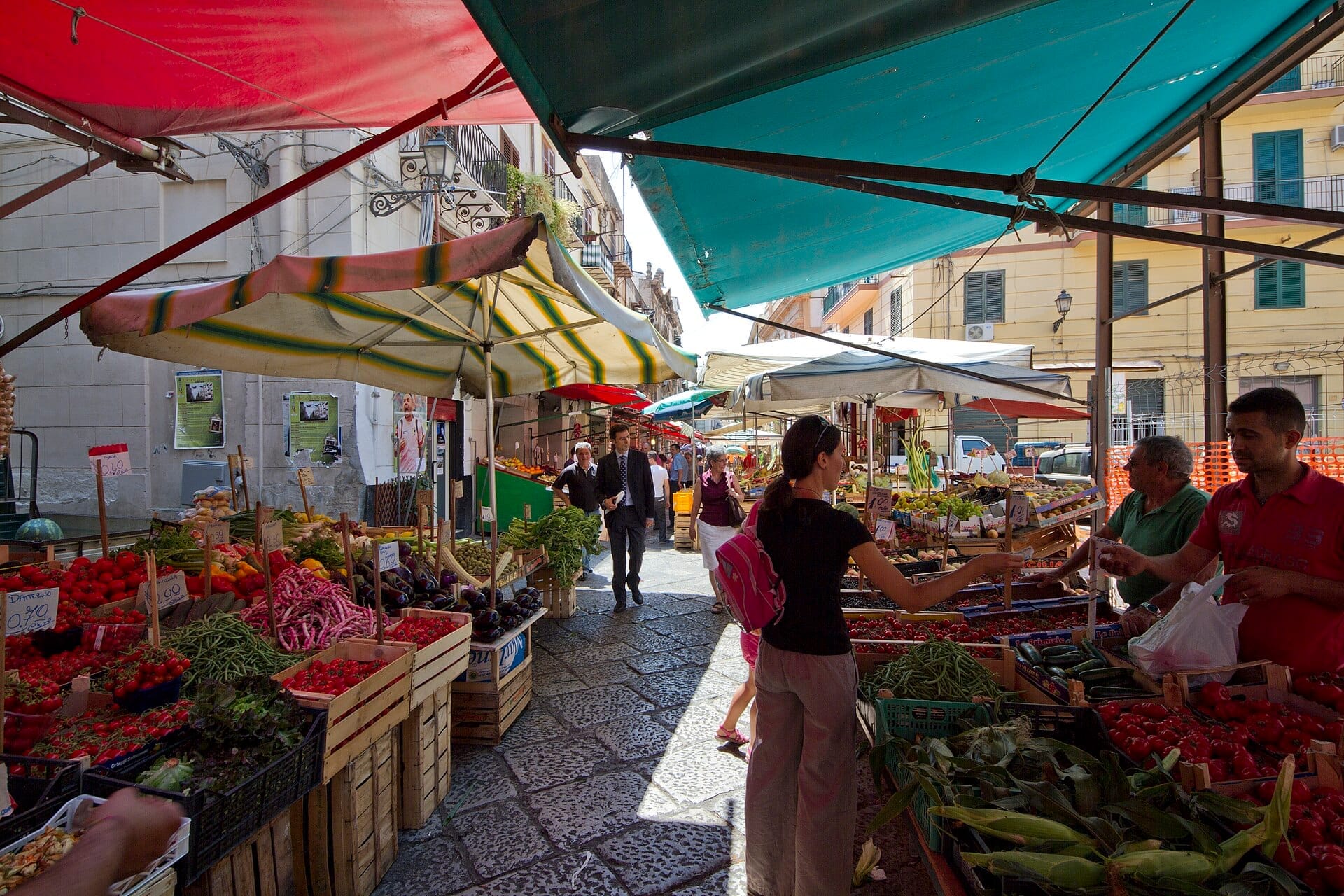 Stände mit frischen Produkten unter bunten Markisen auf dem Vucciria-Markt in Palermo, mit Einkäufern, die auf dem lebhaften Platz Obst und Gemüse begutachten.