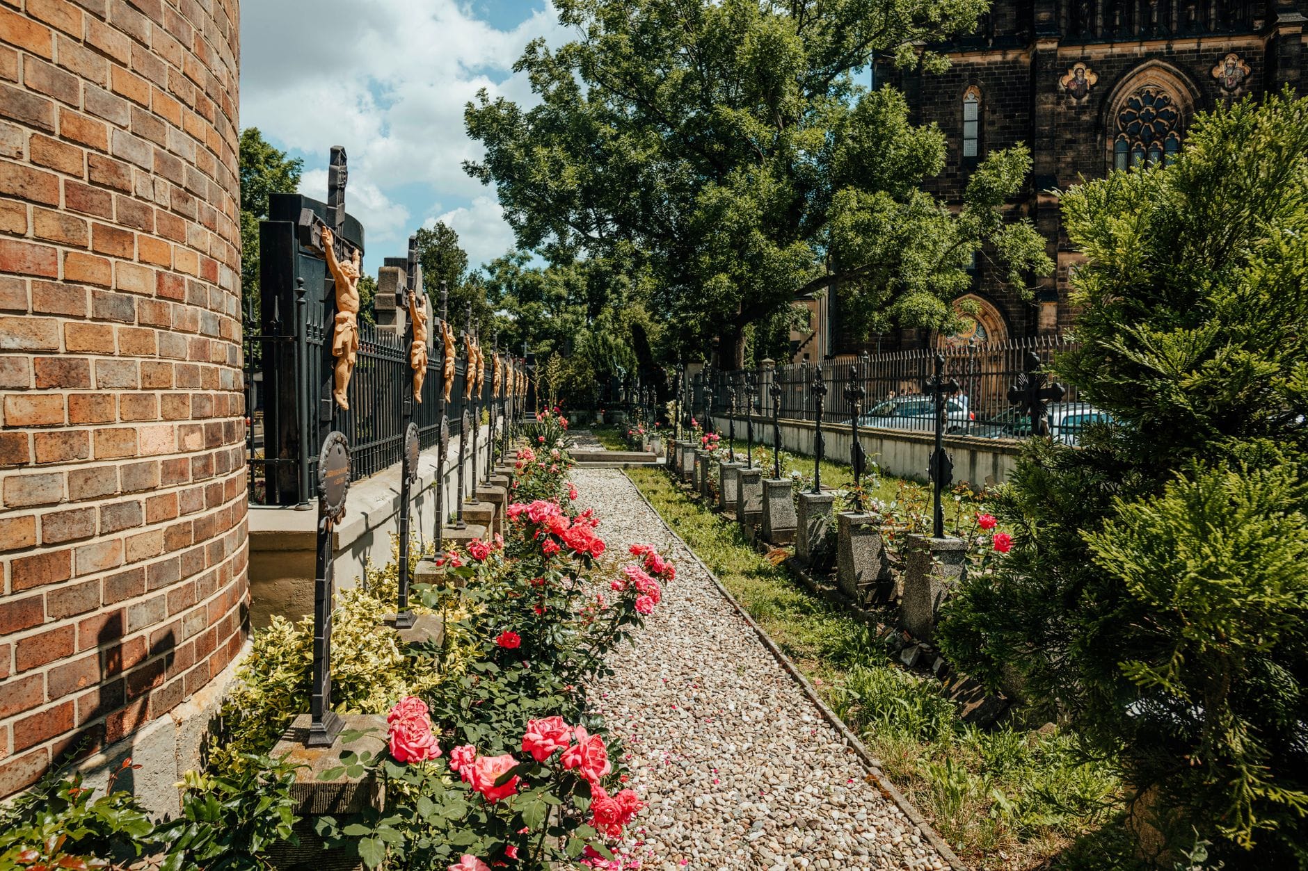 Mit kunstvollen Grabsteinen und bunten Blumen gesäumter Weg auf dem Vyšehrad-Friedhof, im Hintergrund üppiges Grün und die historische Kirchenarchitektur unter blauem Himmel.