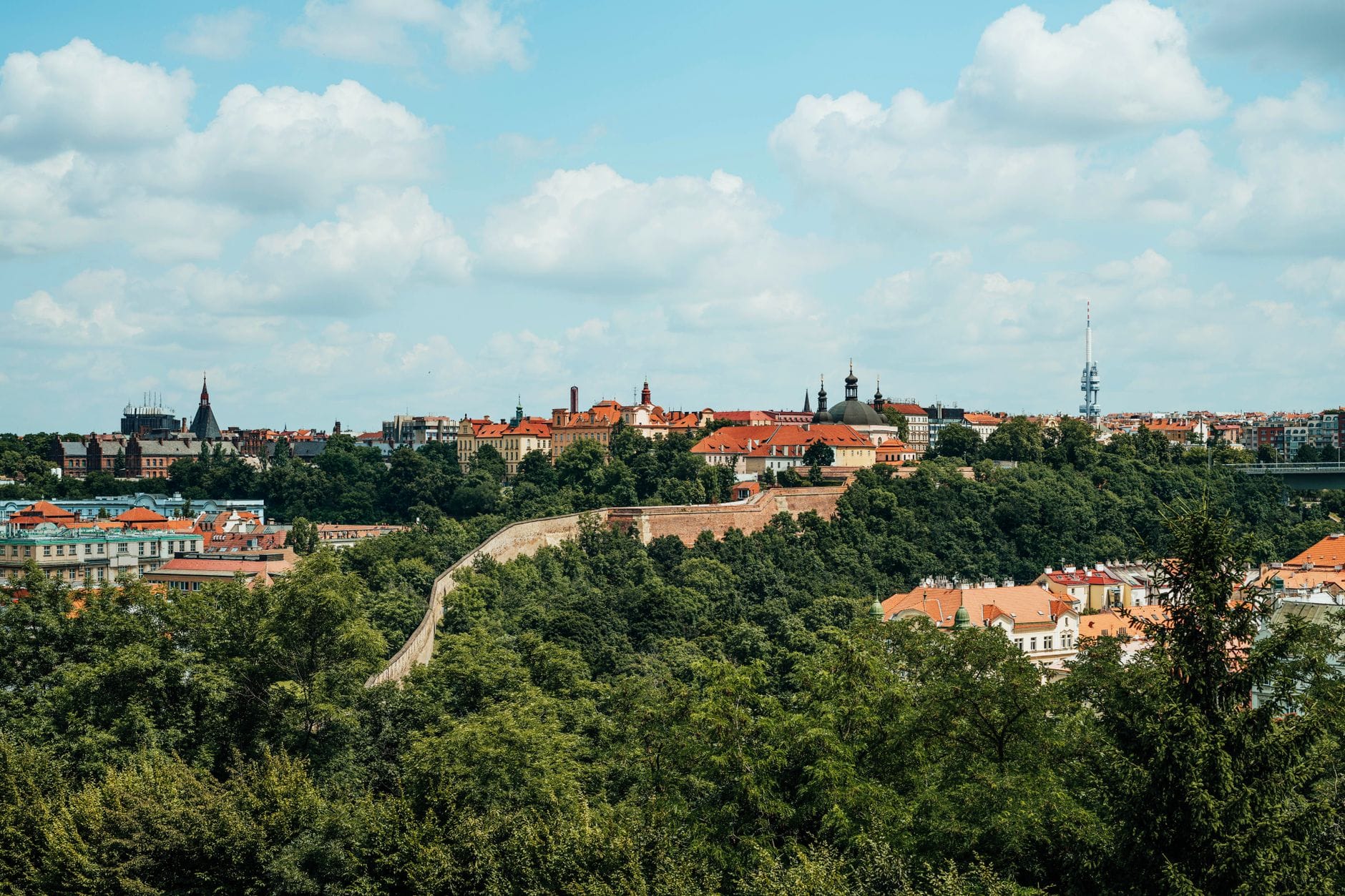 Weitläufige Landschaftsansicht der Festung Vyšehrad auf einem grünen Hügel mit ihren markanten Türmen über den Bäumen, mit Blick auf Prag unter einem leicht bewölkten Himmel.