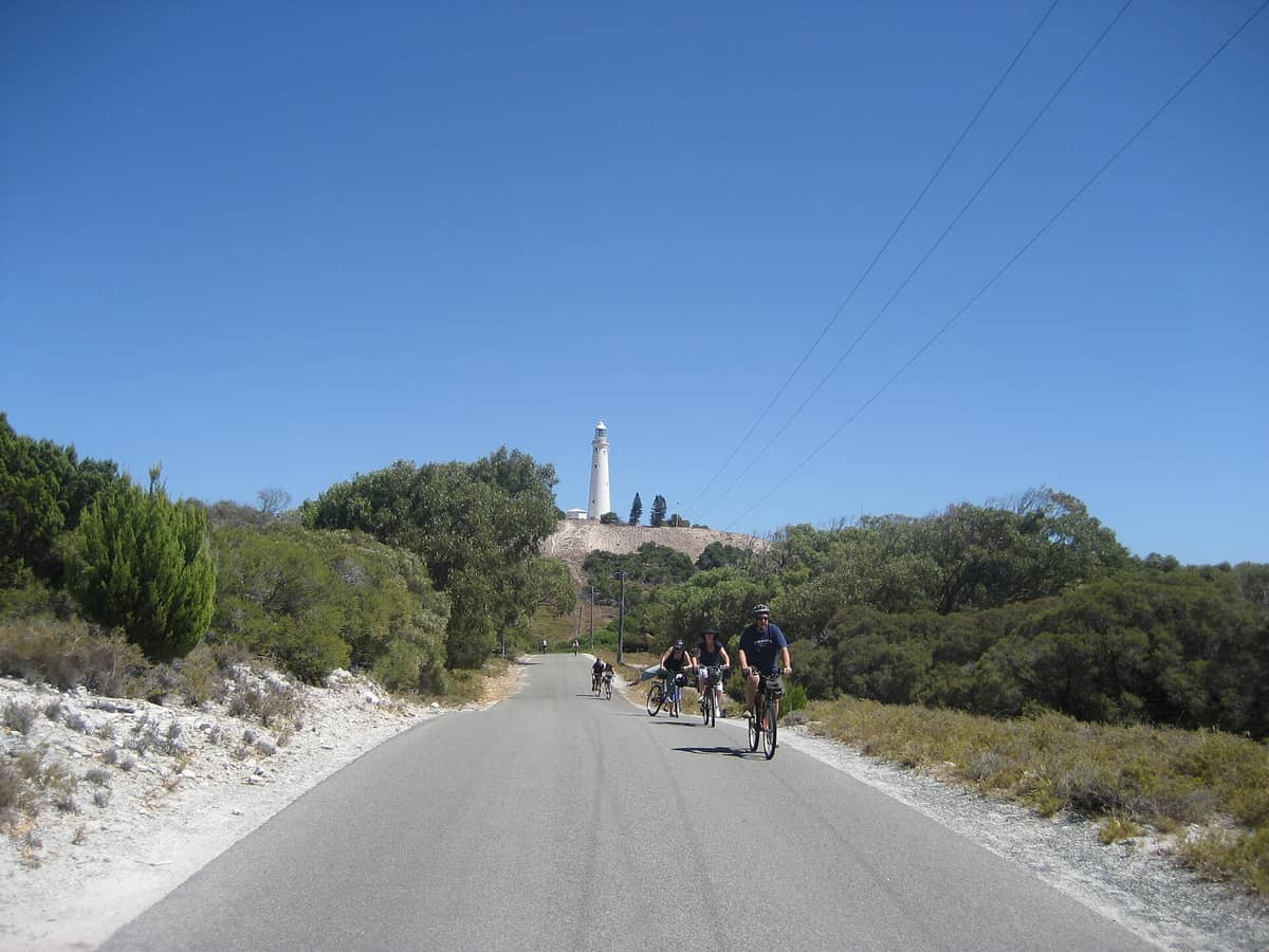 View looking up the road to Wadjemup Hill on Rottnest Island, with Wadjemup Lighthouse prominently on the hilltop under a wide blue sky.