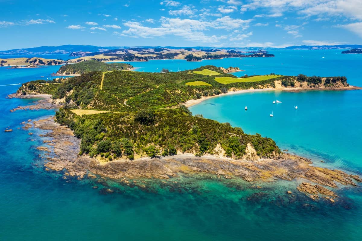 Luftaufnahme von Waiheke Island mit türkisfarbenem Wasser, sattgrünen Hügeln, geschützten Buchten und vor der malerischen Küste ankernden Booten unter strahlend blauem Himmel.