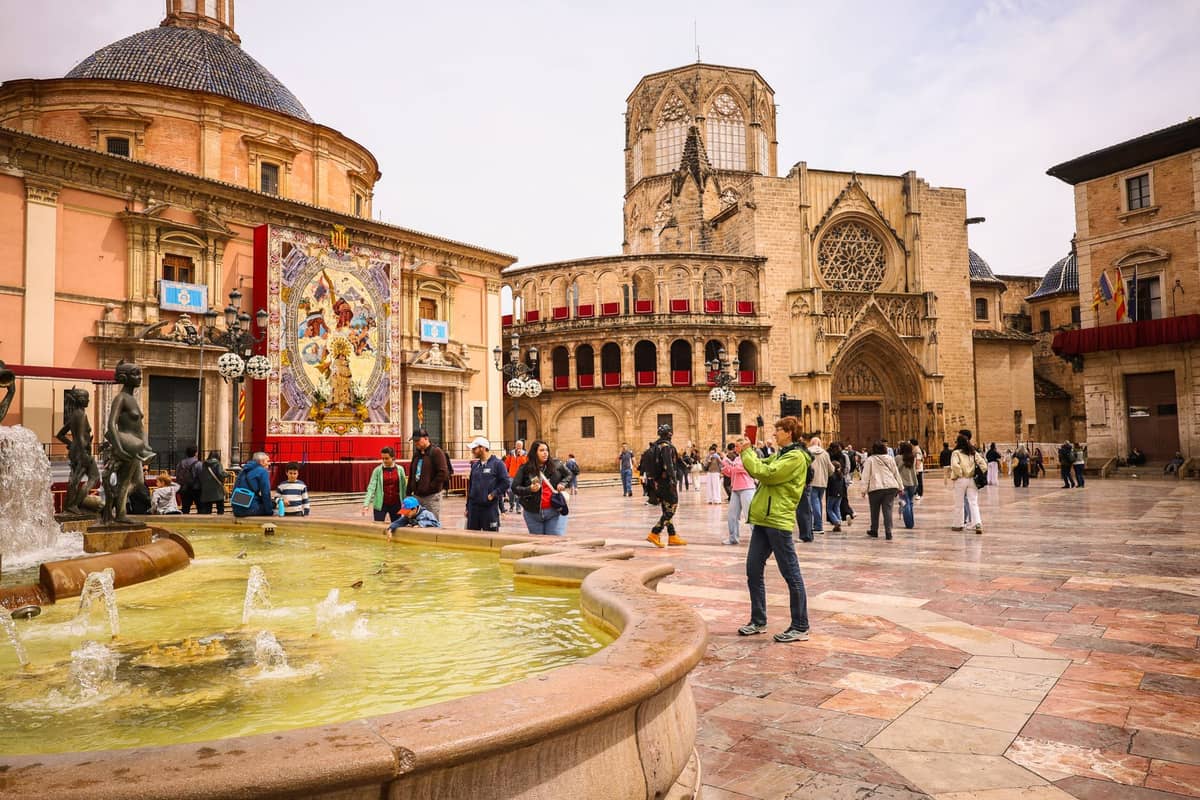 Plaza de la Virgen nella Città Vecchia di Valencia, con la cattedrale, la Basílica de la Virgen, la fontana e i visitatori che esplorano la storica piazza in una giornata nuvolosa.