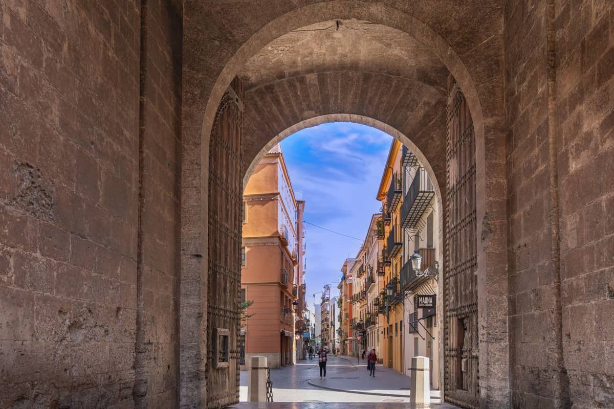 Vista a través de un arco de piedra hacia una calle peatonal flanqueada de edificios históricos de Valencia, con algunas personas paseando.