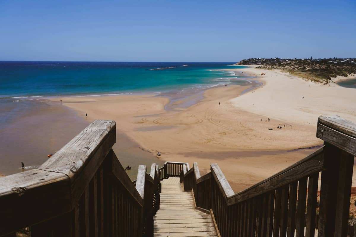 Wooden stairs descend to a wide sandy beach with sweeping ocean views and a distant Adelaide suburb under a bright blue sky.
