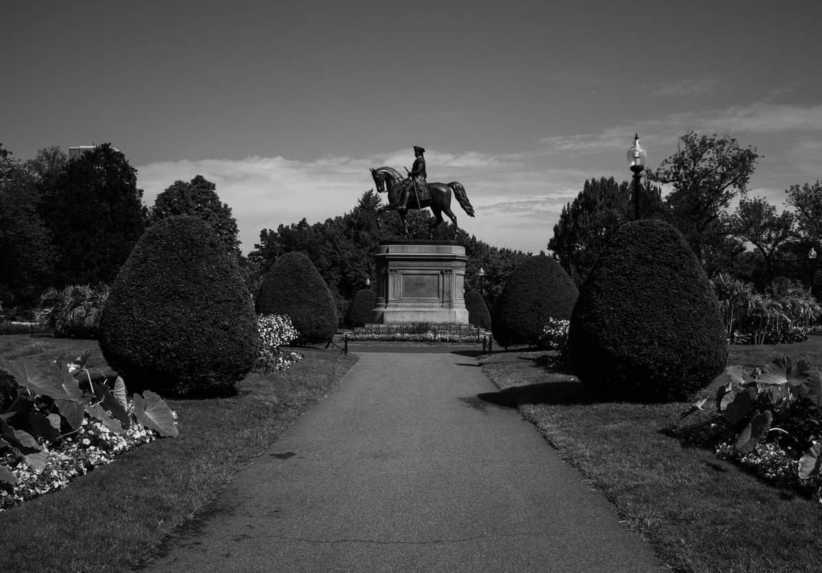 Una estatua de un hombre a caballo a lo largo de un camino bordeado de jardines en el Public Garden de Boston bajo un cielo despejado.