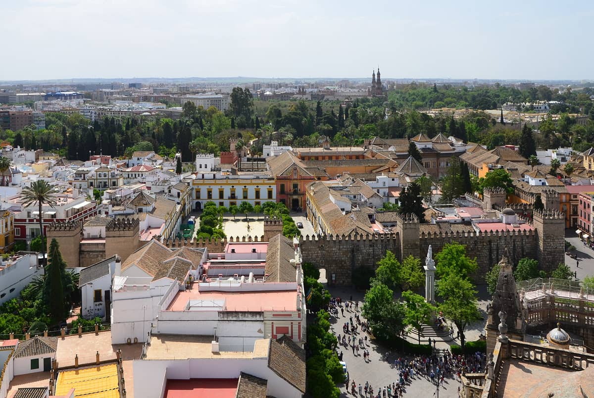 มุมมองทางอากาศของใจกลางเมืองประวัติศาสตร์ของเซบียา รวมถึง Alcázar, Plaza del Triunfo และอาคารโดยรอบพร้อมต้นไม้และผู้คนที่มองเห็นได้ด้านล่าง
