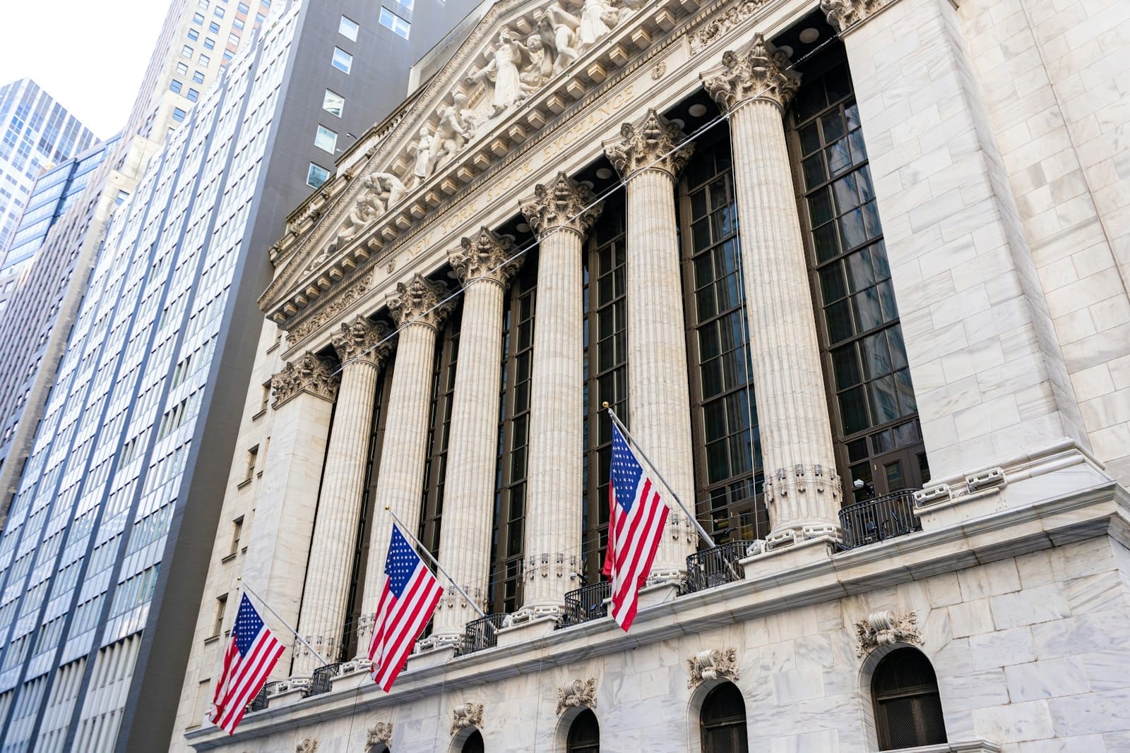 La facciata della New York Stock Exchange su Wall Street, con grandi colonne neoclassiche e tre bandiere americane in una giornata di sole.