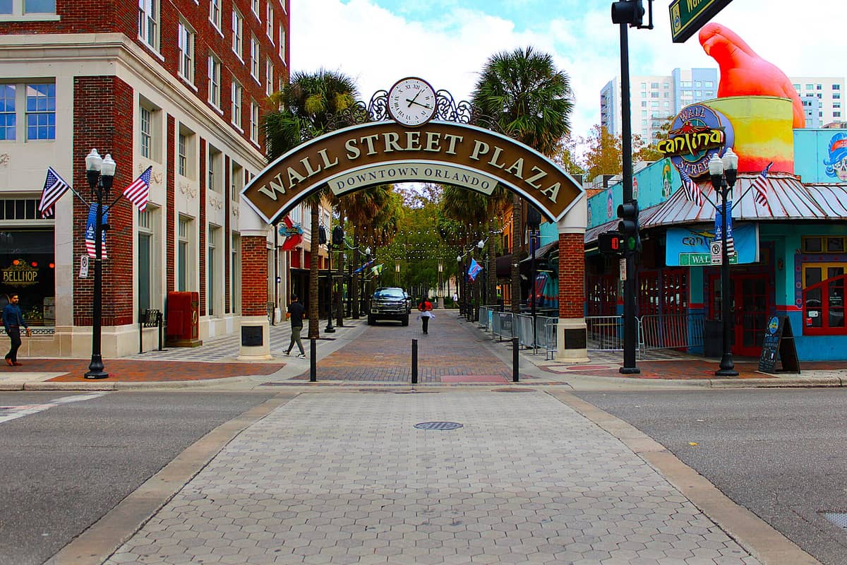 Entrance to Wall Street Plaza in downtown Orlando with an archway sign, colorful building, palm trees, and people walking under string lights.