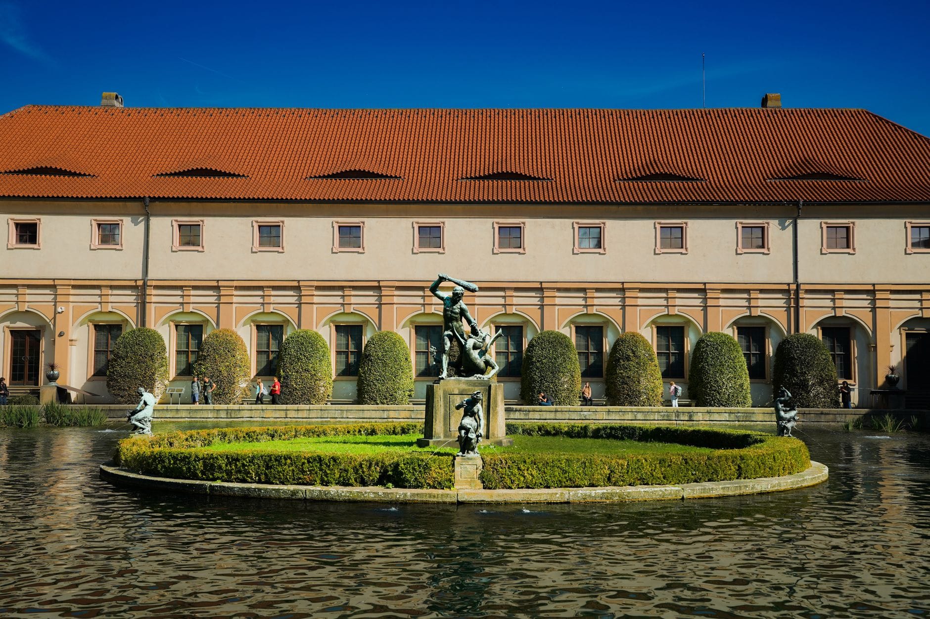 Mittelbrunnen mit Bronzestatuen und gepflegten Hecken im Wallenstein-Garten, historischer Barockpalast im Hintergrund unter strahlend blauem Himmel.