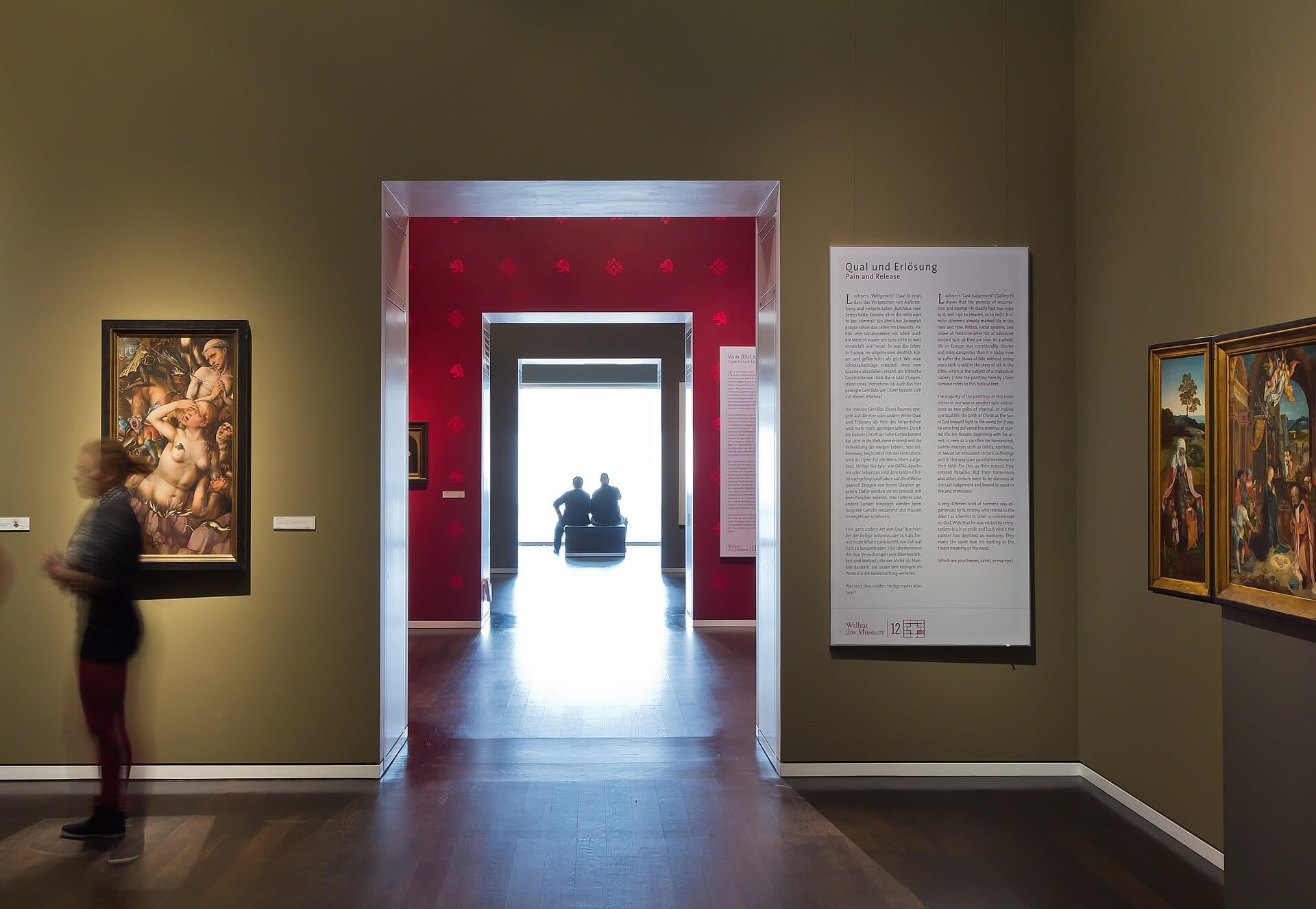 ภายในแกลเลอรีของ Wallraf-Richartz Museum พร้อมภาพวาดยุคกลาง ผู้เยี่ยมชมที่เบลอกำลังเดิน และสองคนนั่งอยู่ใกล้หน้าต่างที่สว่าง