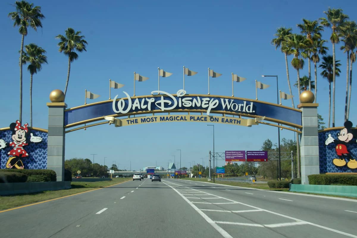 The iconic Walt Disney World entrance arch with colorful flags and images of Mickey and Minnie, welcoming visitors by car on a sunny day.