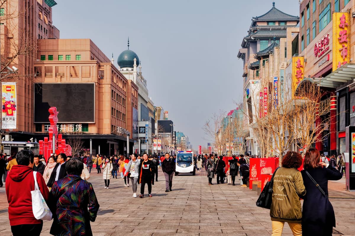 Weitläufige Fußgängerperspektive der Wangfujing-Straße mit Menschenmassen, modernen Gebäuden und traditionellen chinesischen Architekturelementen an einem klaren Tag in Peking.