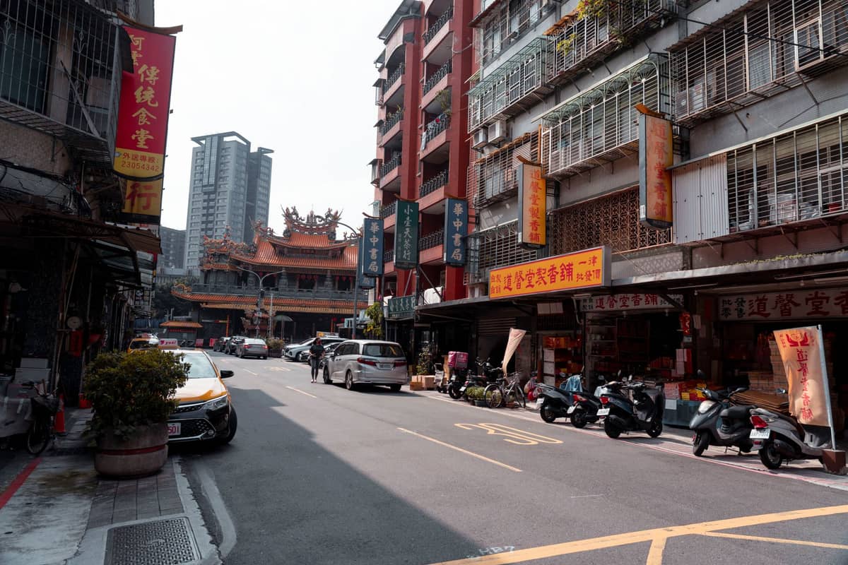 Straßenszene im Wanhua-Viertel mit Motorrollern, Autos und Geschäften – im Hintergrund ist zwischen den Stadtgebäuden der prächtige Longshan-Tempel zu sehen.