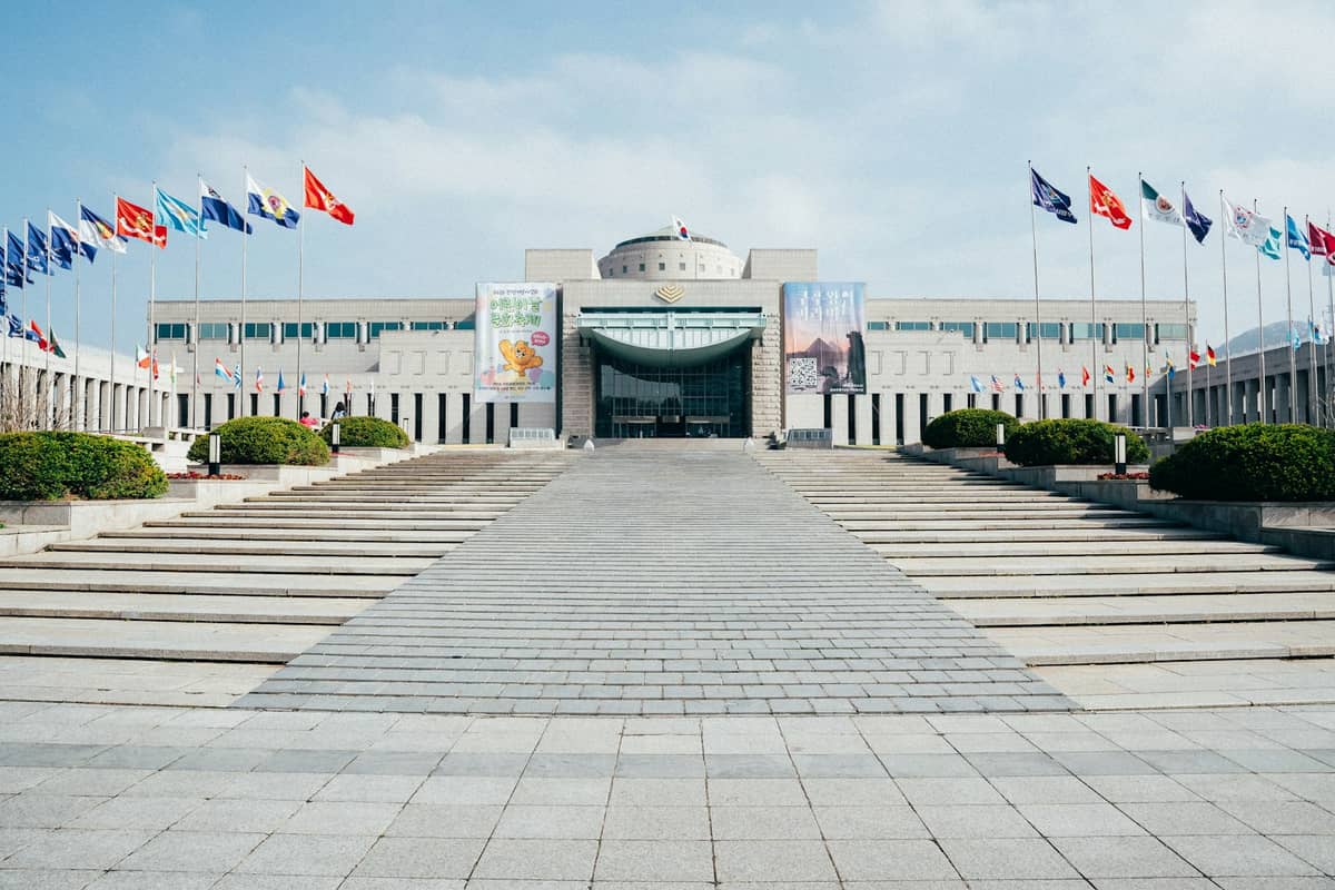 Vista ampla da entrada principal do Memorial de Guerra da Coreia em Seul, com vários mastros de bandeira e uma grande escadaria que leva ao edifício em um dia de céu aberto.