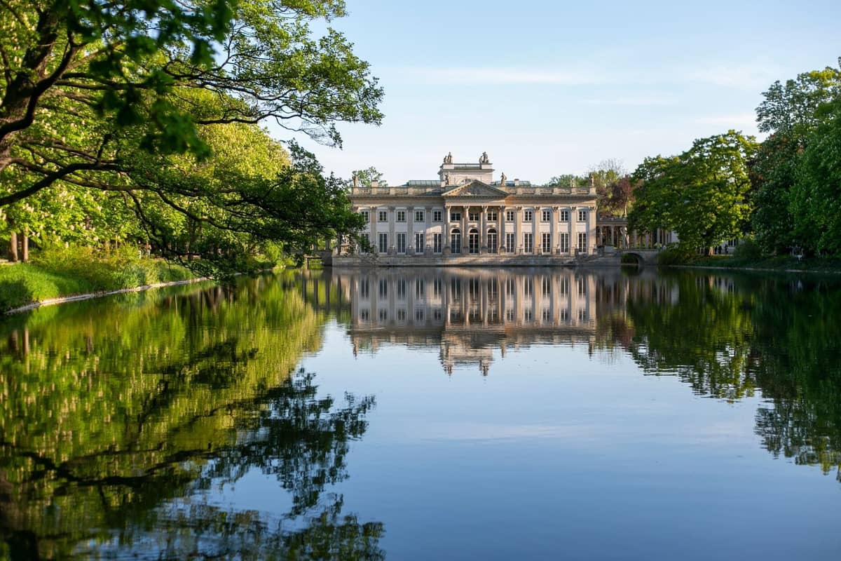 Ein prächtiger neoklassizistischer Palast, der sich an einem klaren Tag im ruhigen Teich des Königlichen Łazienki-Parks in Warschau spiegelt, umgeben von üppigen grünen Bäumen.