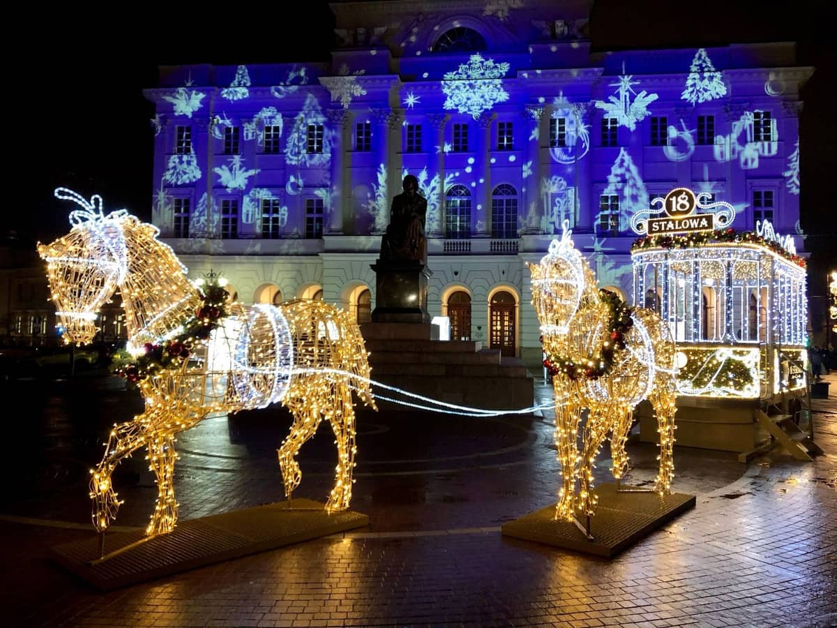 Scène nocturne dans le centre-ville de Varsovie avec un affichage lumineux de Noël représentant un cheval et une calèche illuminés devant un grand bâtiment historique, projections festives et décorations.