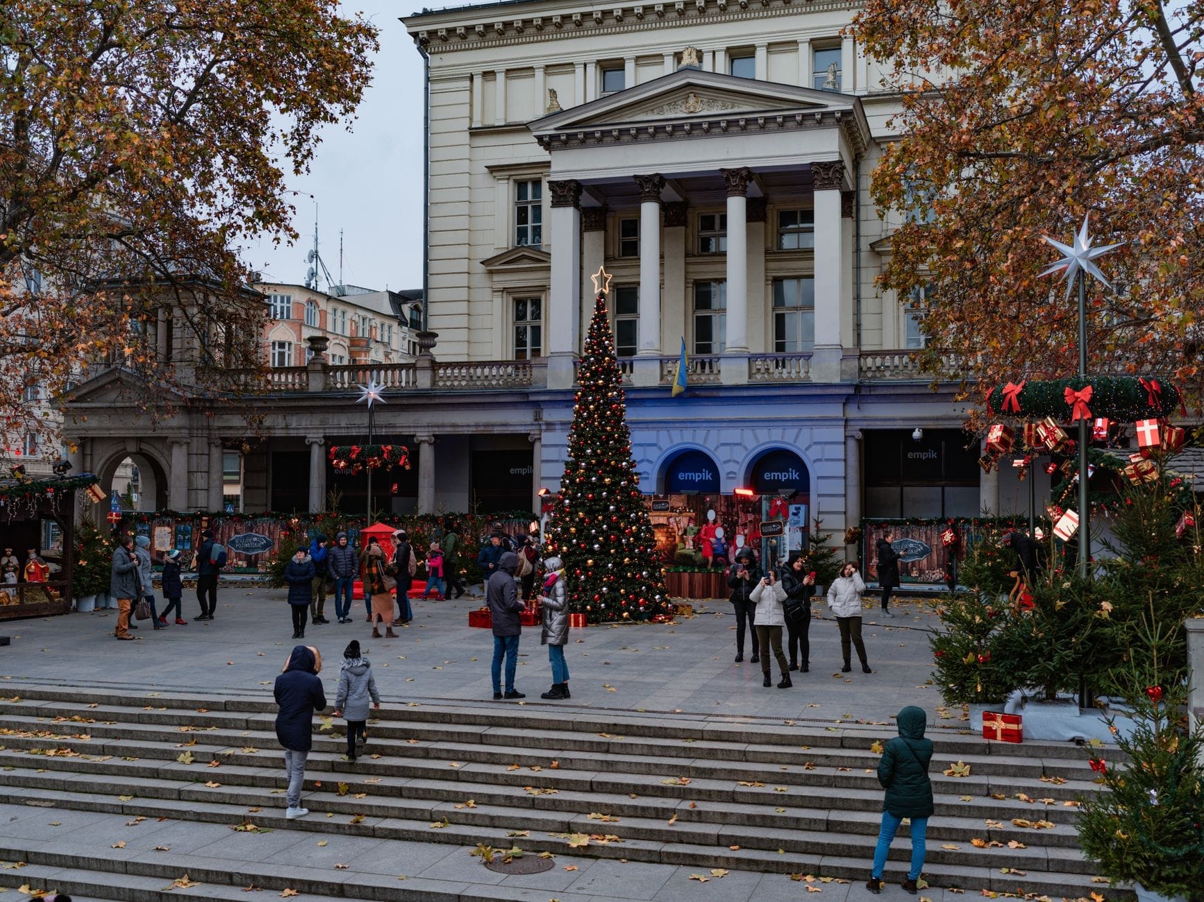 Pessoas curtindo o mercado de Natal de Varsóvia em frente a um imponente edifício neoclássico, com uma grande árvore de Natal decorada e barracas festivas em uma praça ao ar livre.