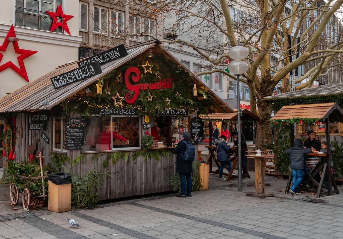 Chalet en bois de marché de Noël sur une place de ville avec des personnes commandant de la nourriture et des boissons festives, des tables en plein air et des décorations de fête.