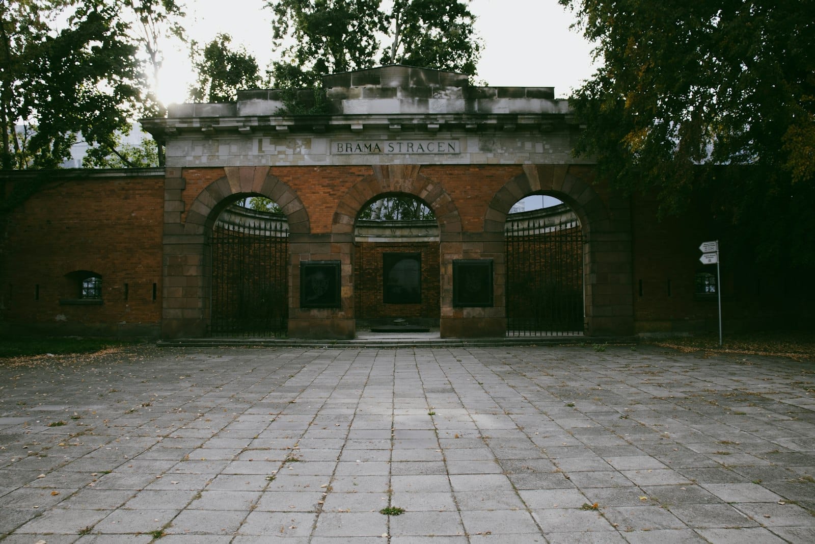 ทางเข้า Brama Straceń ของ Warsaw Citadel แสดงให้เห็นประตูโค้งก่ออิฐพร้อมบานประตูเหล็กดัด ล้อมรอบด้วยต้นไม้ในยามพลบค่ำ